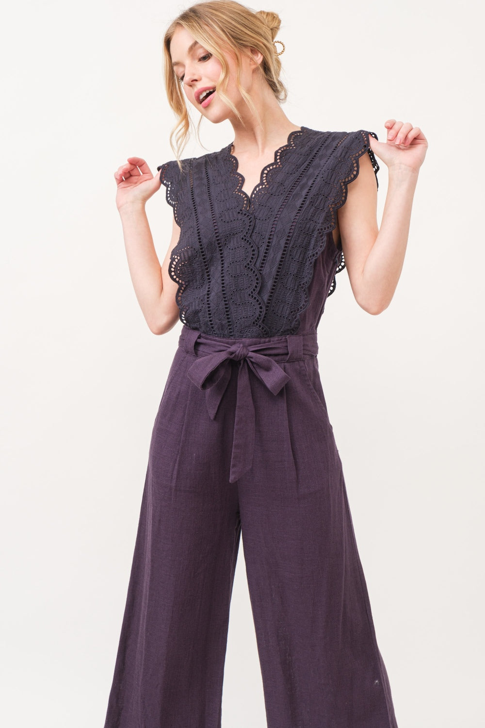 A person in an elegant and fashionable And The Why Laced Surplice Tie Waist Jumpsuit with wide-leg pants stands against a plain background, smiling with hands on hips.