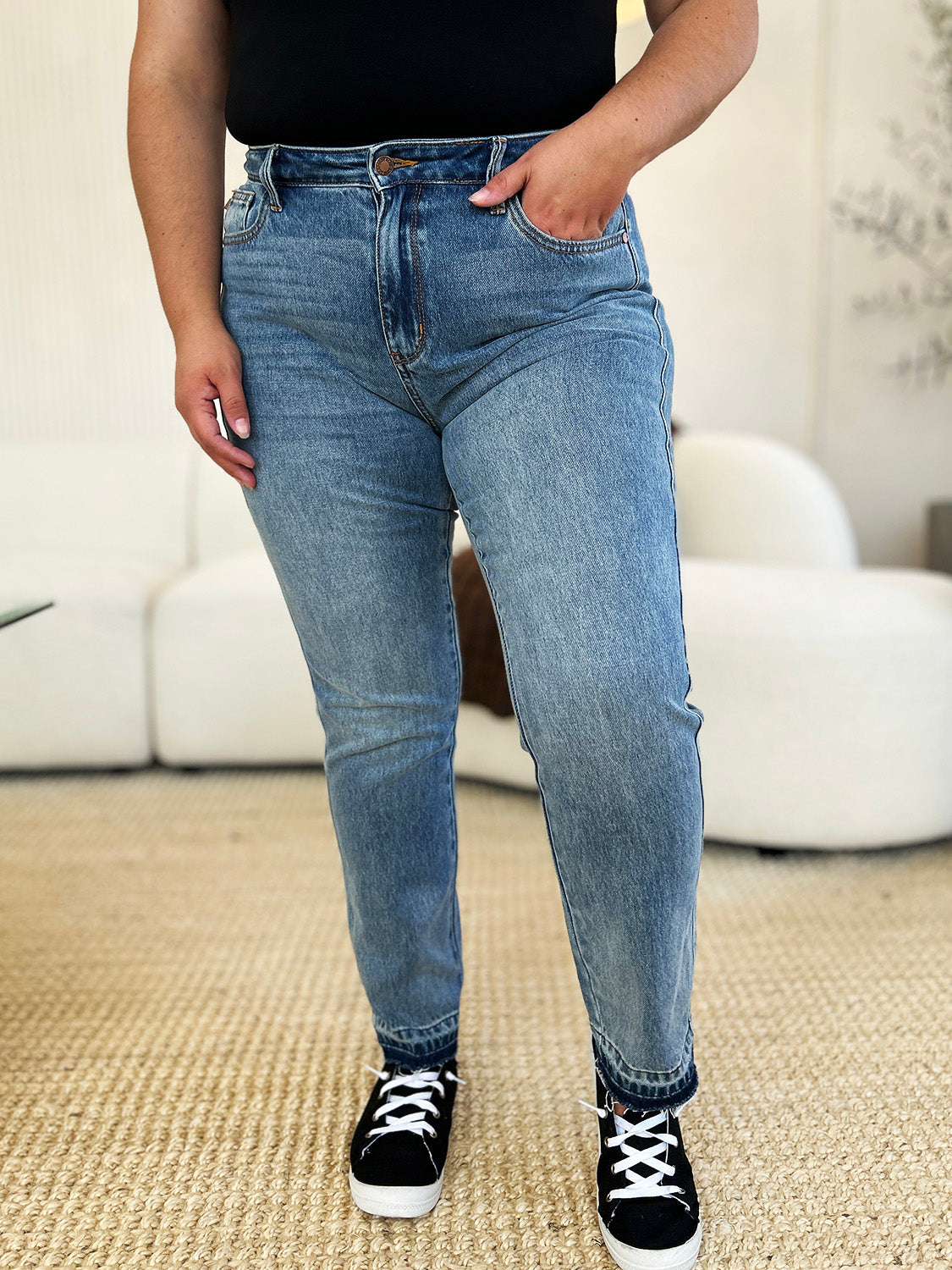 Person wearing a black top, Judy Blue Full Size Mid Rise Rigid Magic Release Hem Jeans, and black sneakers with white laces, standing on a beige carpet with hands in pockets.