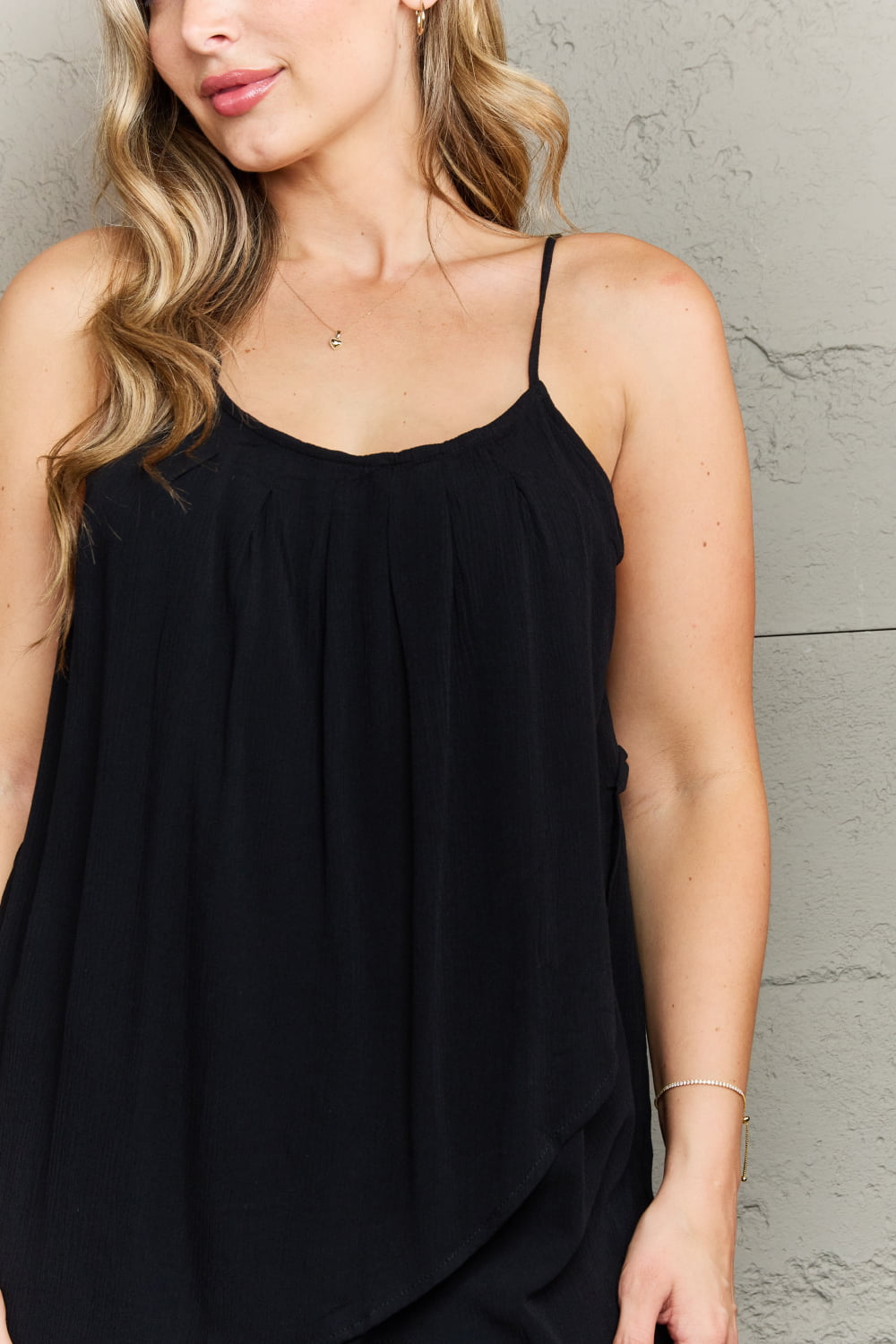 A person with long hair in the Ninexis Hang Loose Tulip Hem Cami Top and blue jeans stands against a textured gray wall, smiling at the camera. The tulip wrap hem of the top adds a stylish touch to her outfit.