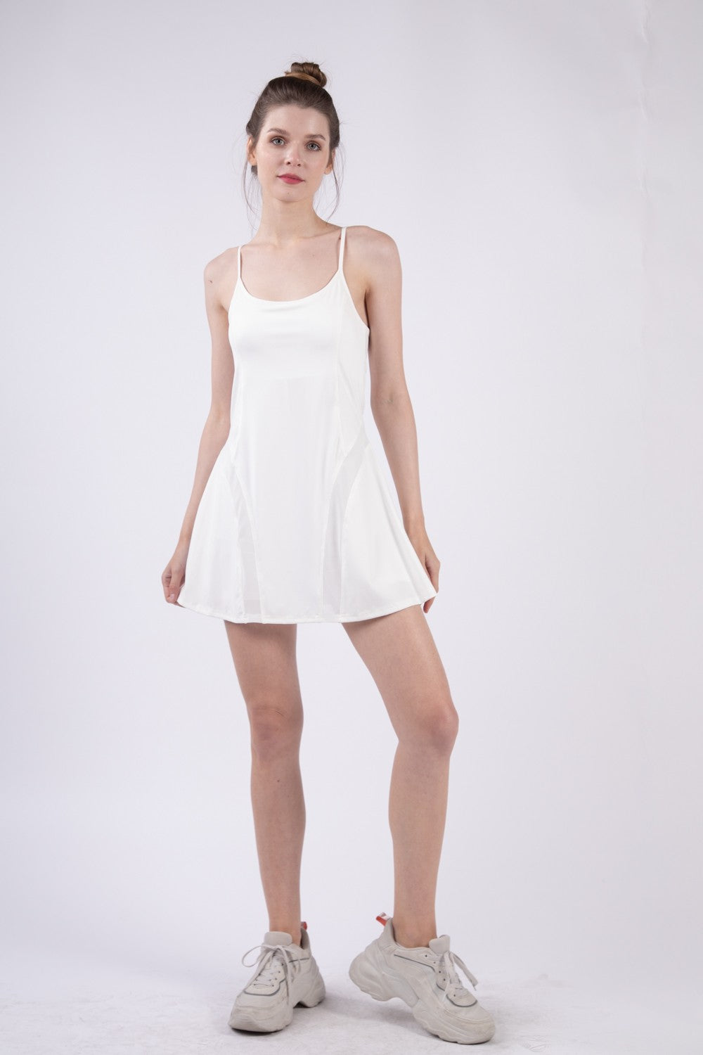 A woman stands against a plain backdrop, wearing the VERY J Mesh Detail Tennis Mini Active Dress with Shorts Inside and light-colored sneakers. She has her hair tied back and is looking directly at the camera, exuding an effortlessly sporty vibe in her activewear dress.