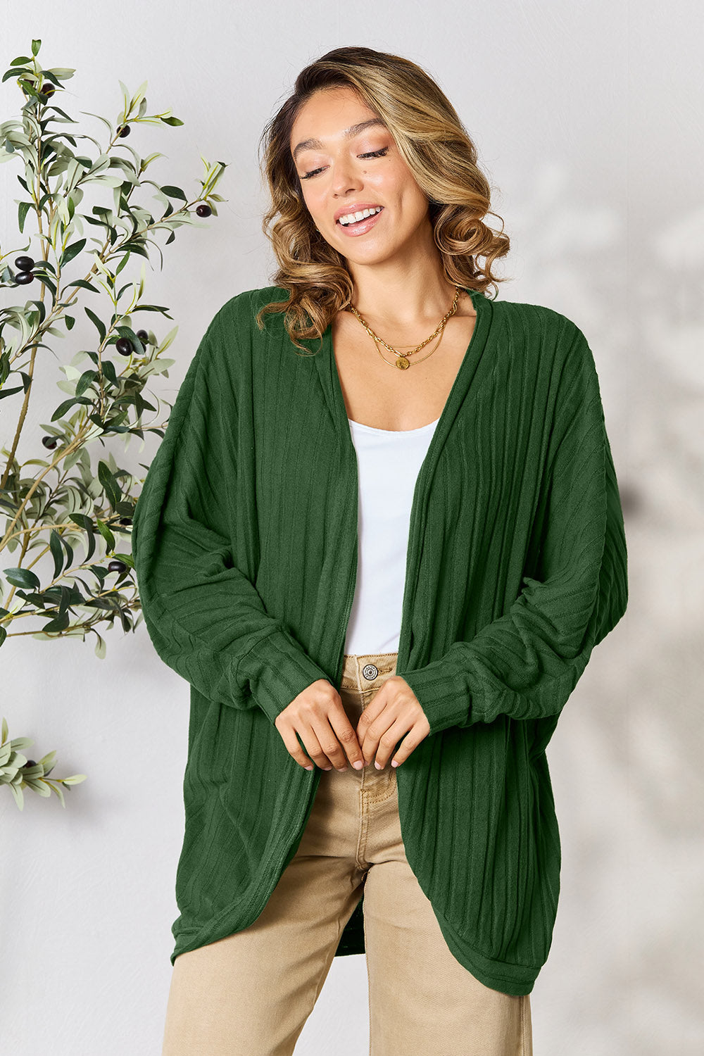 A person with wavy hair, wearing the Basic Bae Full Size Ribbed Cocoon Cardigan over a white top and beige pants, stands in front of a light-colored background with a leafy plant, showcasing an effortlessly basic style.