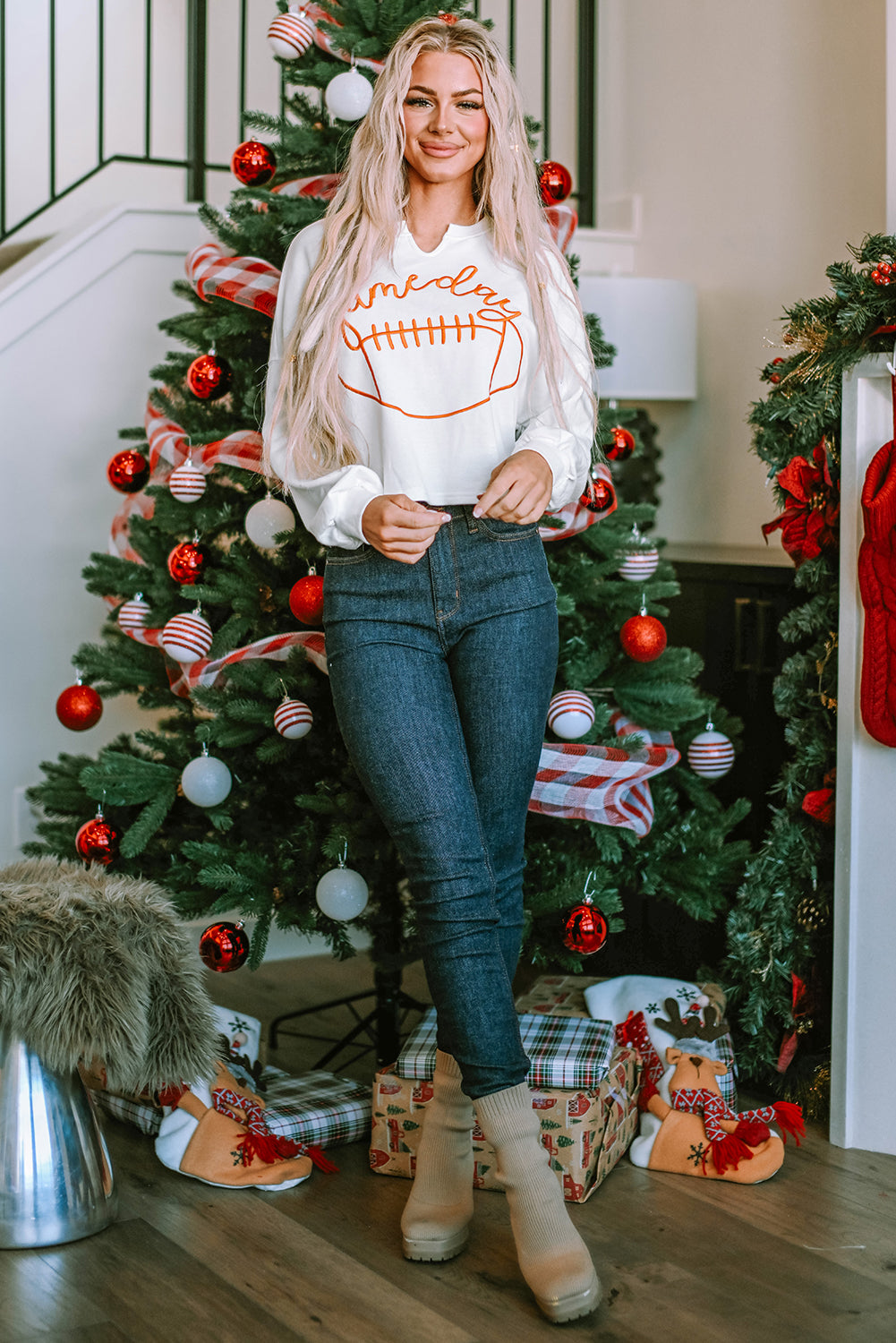 A woman with long blonde hair dressed in a chic White Game Day Lettering Rugby Football Notched Neck Sweatshirt and blue jeans stands near a Christmas tree adorned with red and white ornaments and ribbon.