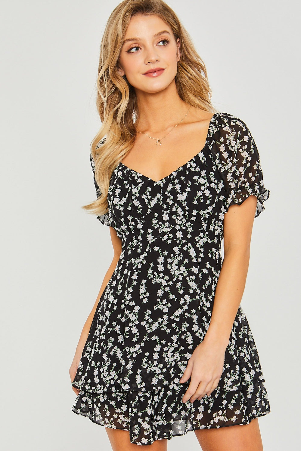 A woman is showcasing summer fashion in a short black floral romper from Love Tree, standing against a plain light background.