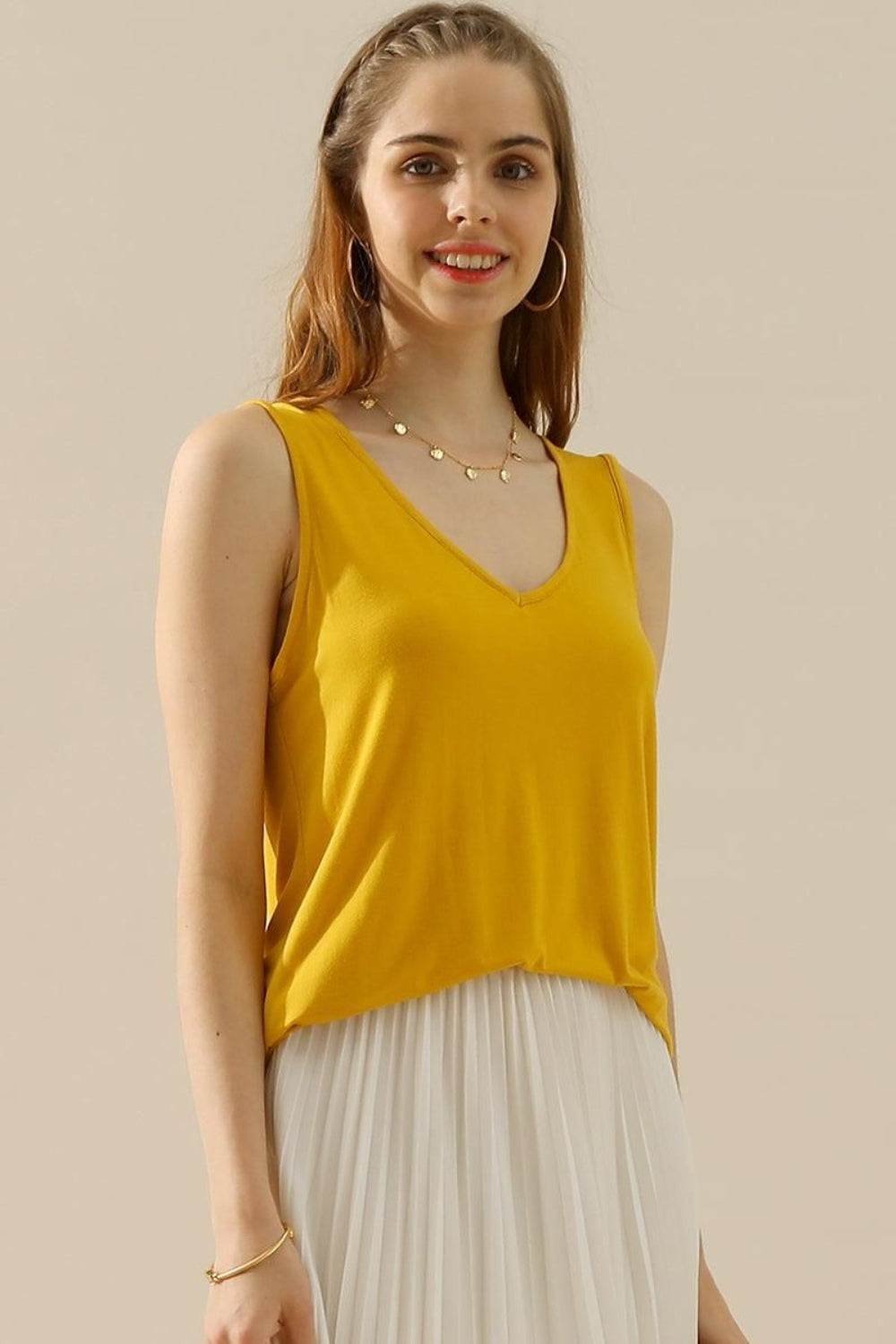 A woman in a stylish and versatile Ninexis Full Size V-Neck Curved Hem Tank and white pleated skirt poses while holding her handbag.