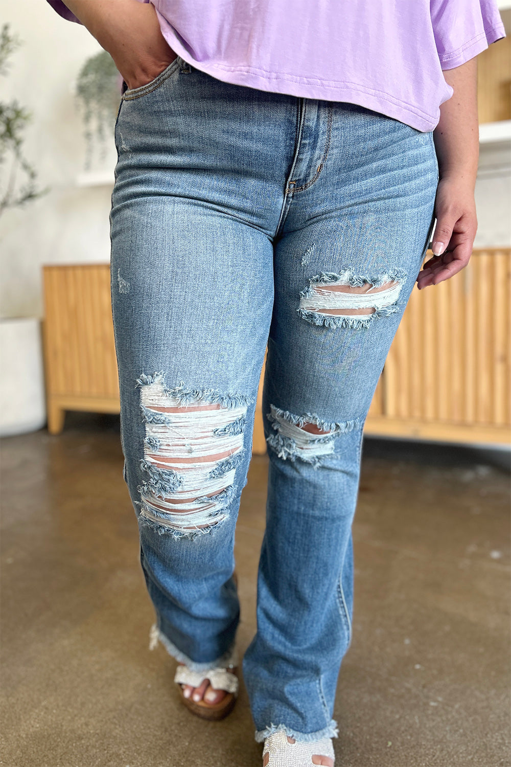 A person wearing Judy Blue Full Size Distressed Raw Hem Bootcut Jeans and a light purple top stands on a polished floor with a wooden cabinet in the background.