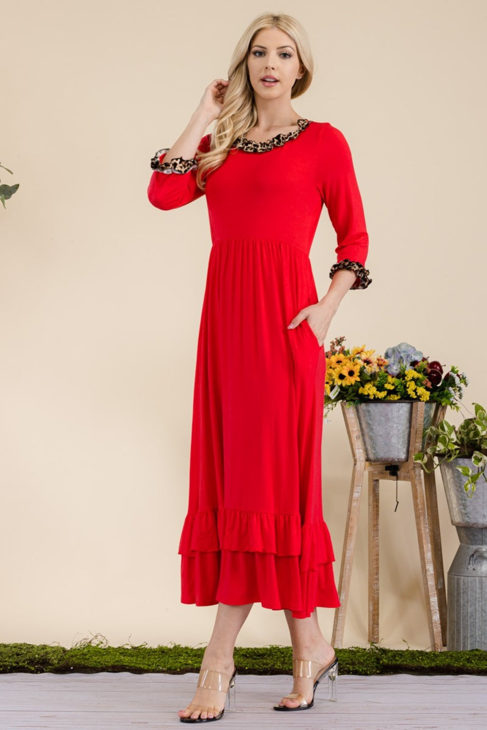 A woman wearing the Celeste Full Size Leopard Ruffle Detail Midi Dress stands smiling next to a pot of flowers, making her look perfect for special occasions.