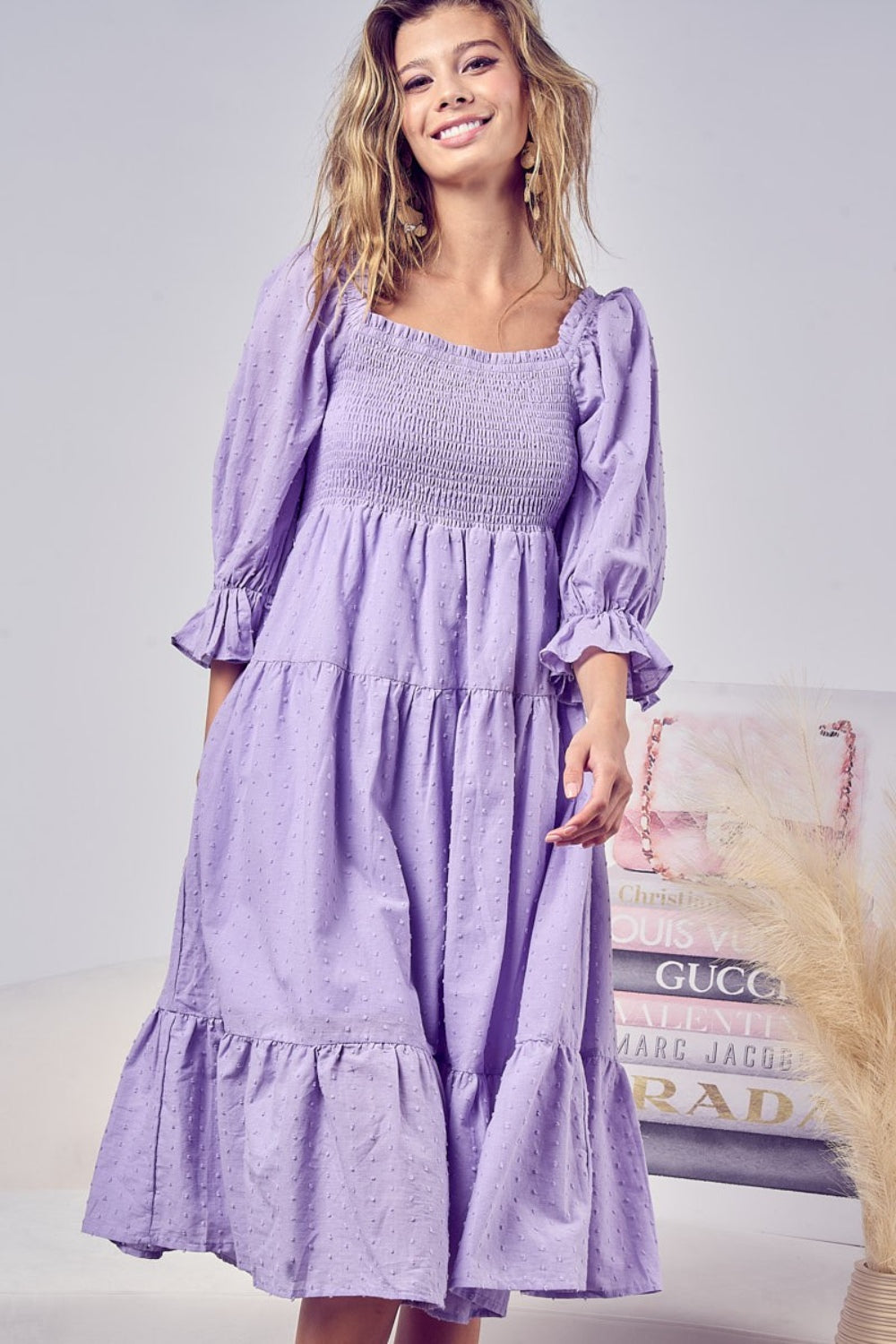 A woman stands smiling in the BiBi Swiss Dot Flounce Sleeve Smocked Tiered Midi Dress, showcasing its lavender hue, long flounce sleeves, tiered skirt, and delicate Swiss dots.