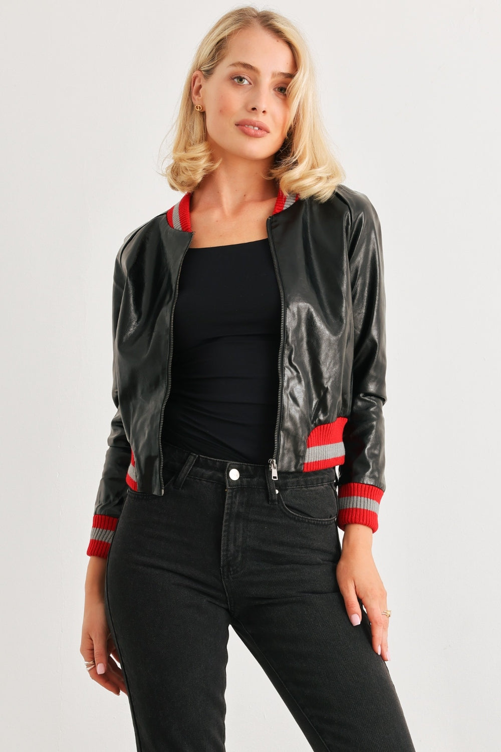 A woman with shoulder-length blonde hair in a black top, black pants, and a trendy Comme PU Leather Baseball Collar Long Sleeve Jacket stands against a plain white background. The jacket features red striped cuffs and a classic baseball collar.