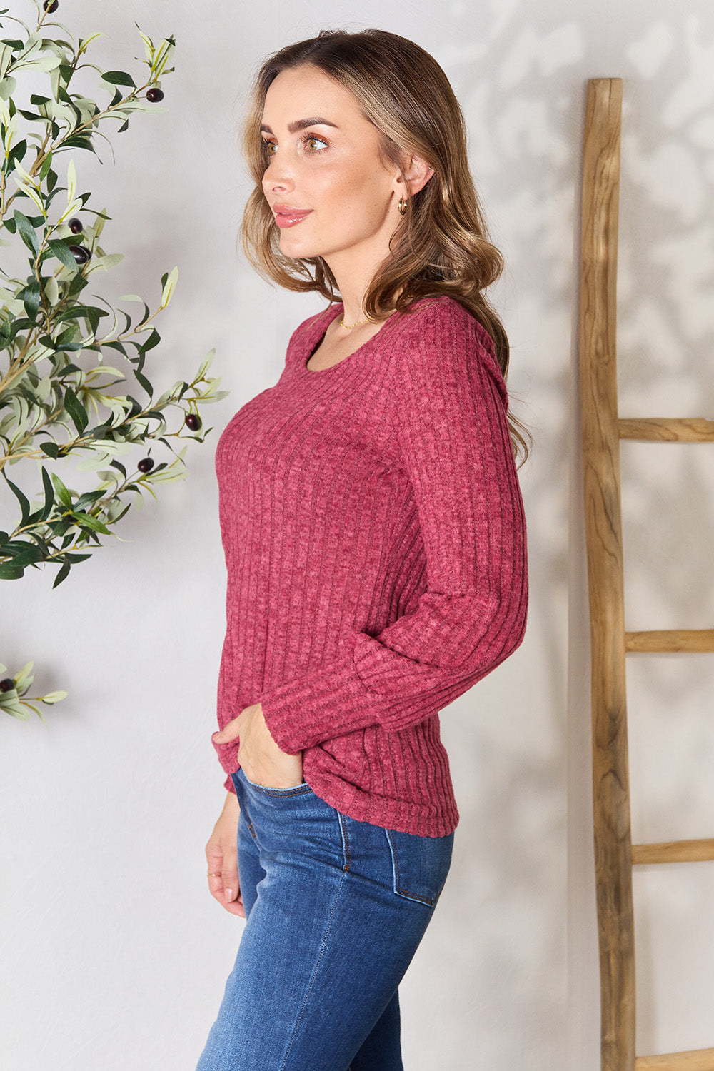 A person in a stylish maroon Ribbed Round Neck Lantern Sleeve Blouse and jeans stands by a wooden ladder, with olive branches in the background.