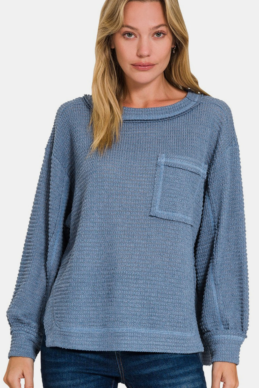 A woman wearing the Zenana Exposed Seam Side Slit Long Sleeve Top, which features a loose fit and textured blue fabric with side slits and a pocket on the left side, stands against a plain, light background. She has blonde hair and is looking directly at the camera.