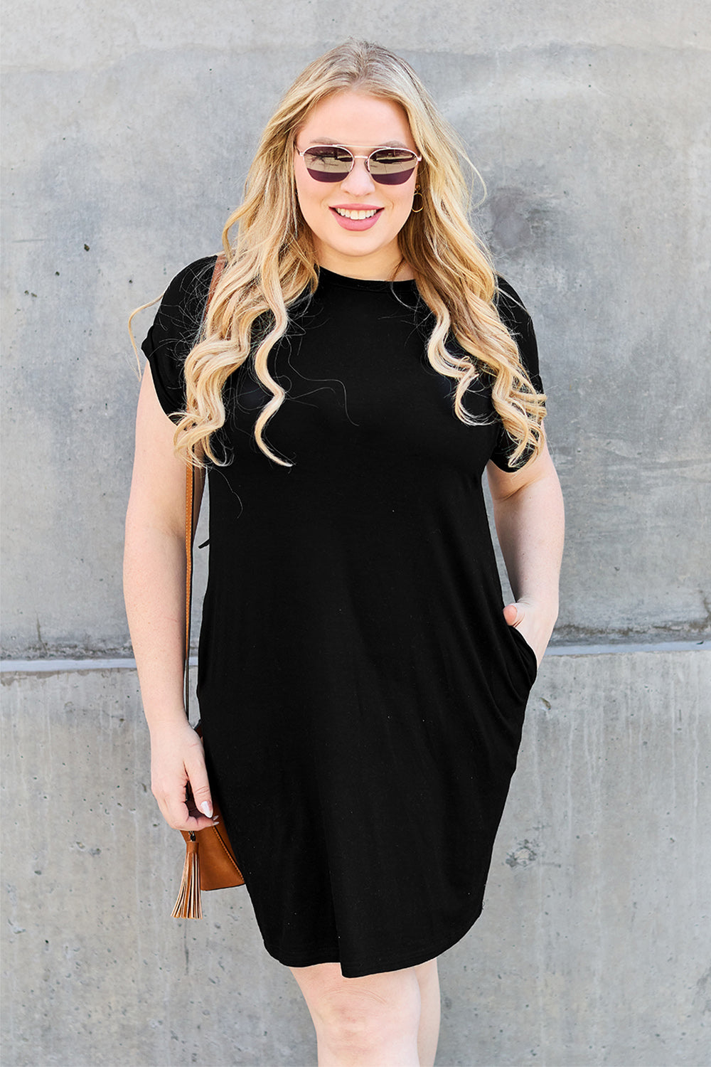 A woman wearing a Basic Bae Bamboo Full Size Round Neck Short Sleeve Dress with Pockets and sunglasses holds the strap of a light-colored backpack while standing against a concrete wall.