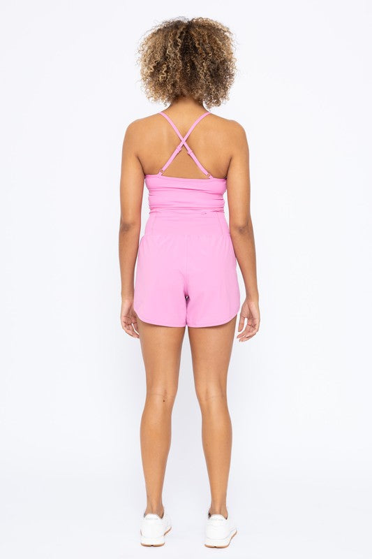 A person in a pink Cross Back Active Romper stands against a white background. They have curly hair and are wearing white sneakers.