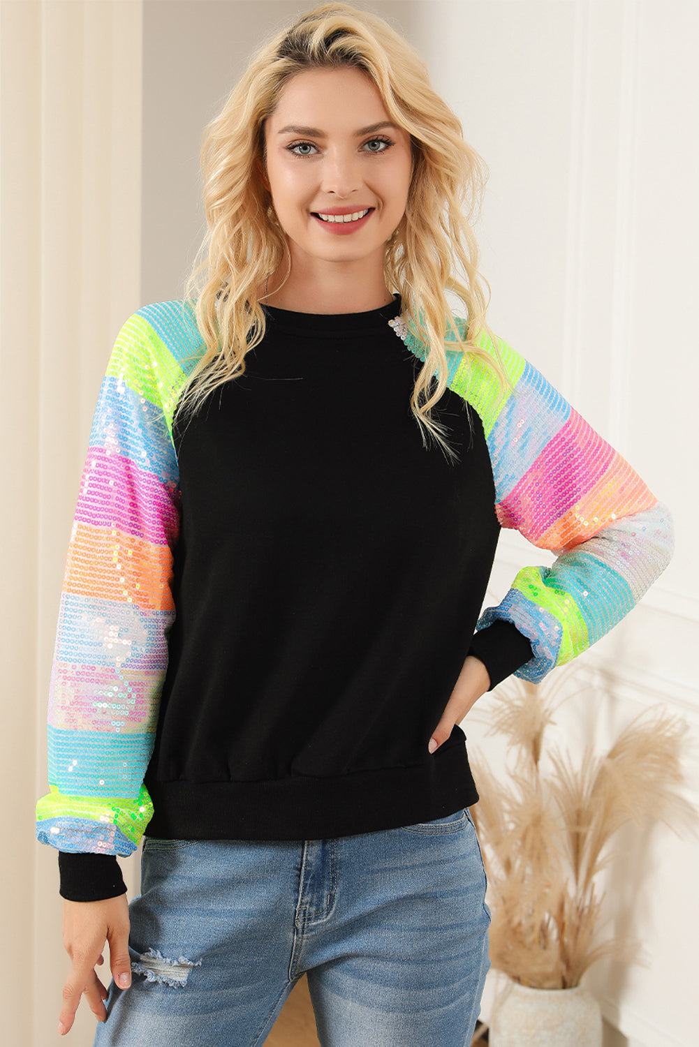 A person with shoulder-length hair is wearing a Black Sequin Color Block Raglan Sleeve Pullover Sweatshirt and blue jeans. They are facing away from the camera in a room with a potted plant.