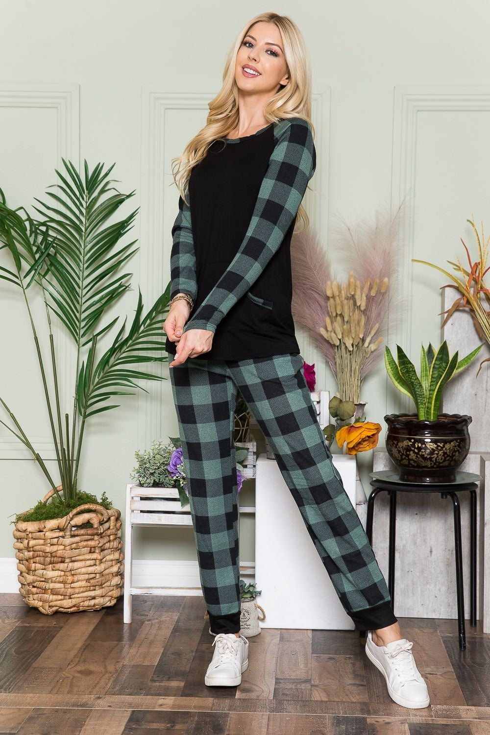 An individual wearing a Celeste Plaid Long Sleeve T-Shirt with Pockets stands indoors on a wooden floor, next to potted plants and a wicker basket.