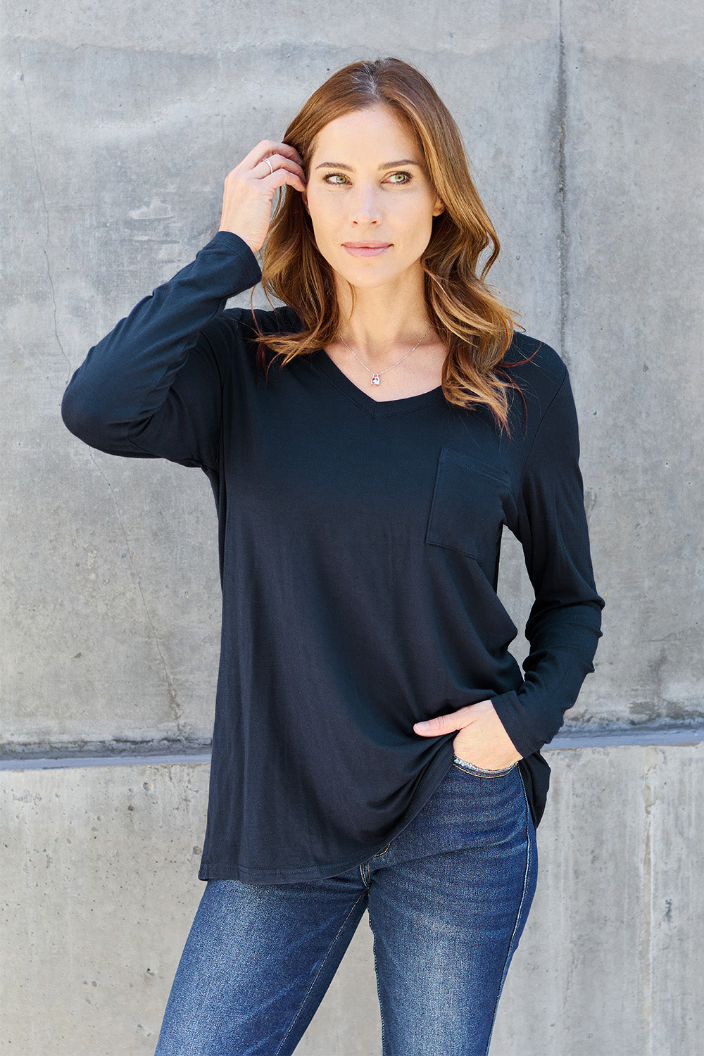 A woman with light brown hair stands against a concrete wall, wearing the Basic Bae Full Size V-Neck Long Sleeve Top in olive green and blue jeans, with a white backpack on her shoulder. Her outfit exudes basic style and is made from polyester, ensuring it’s both comfortable and suitable for machine wash cold. She is looking at the camera.