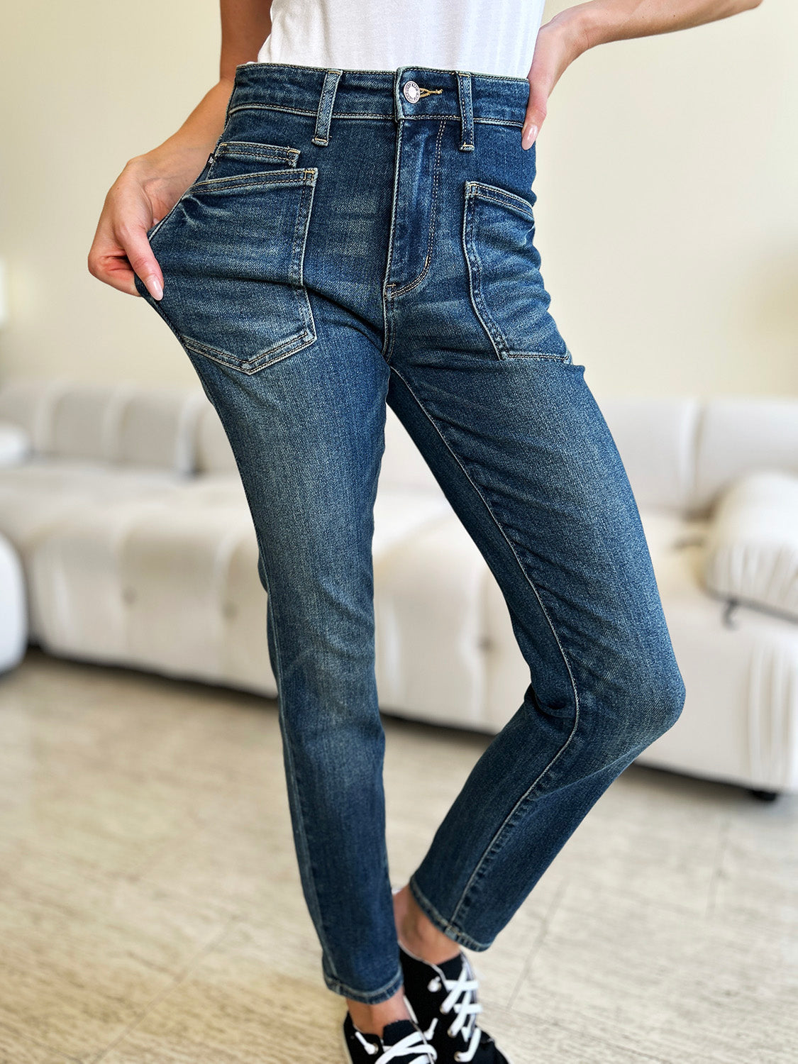 A person wearing Judy Blue Full Size High Waist Skinny Jeans made of stretchy denim fabric stands in a room with light-colored furniture and wooden flooring, paired perfectly with white slip-on shoes.