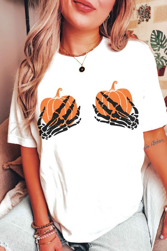 A person wearing the SKELETON HOLDING PUMPKINS Graphic Tee, which features two orange pumpkins and black skeletal hands, is sitting down. With long, wavy hair and accessorized with various bracelets and a necklace, the unisex sizing of the shirt makes it a versatile addition to any wardrobe.