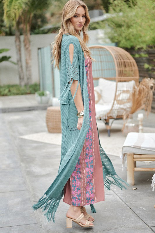 A woman in a green floral dress and a Solid Long Cardigan With Fringe exudes boho-chic vibes as she strolls on a patio, surrounded by outdoor furniture and lush plants.