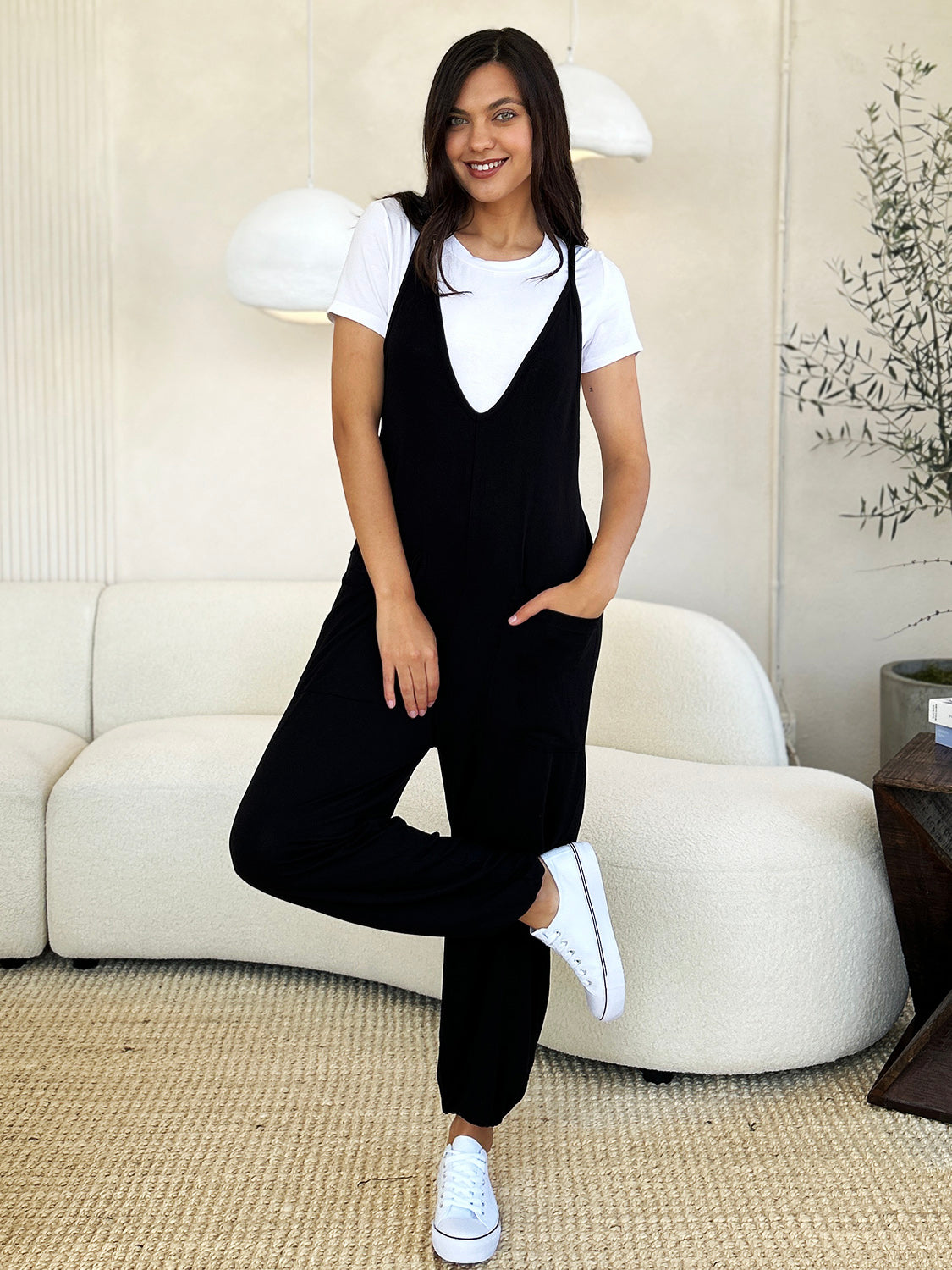 A person wearing the Culture Code Full Size Plunge Sleeveless Jumpsuit with Pockets stands indoors on a beige carpet near a white couch, smiling and holding one hand in their pocket.