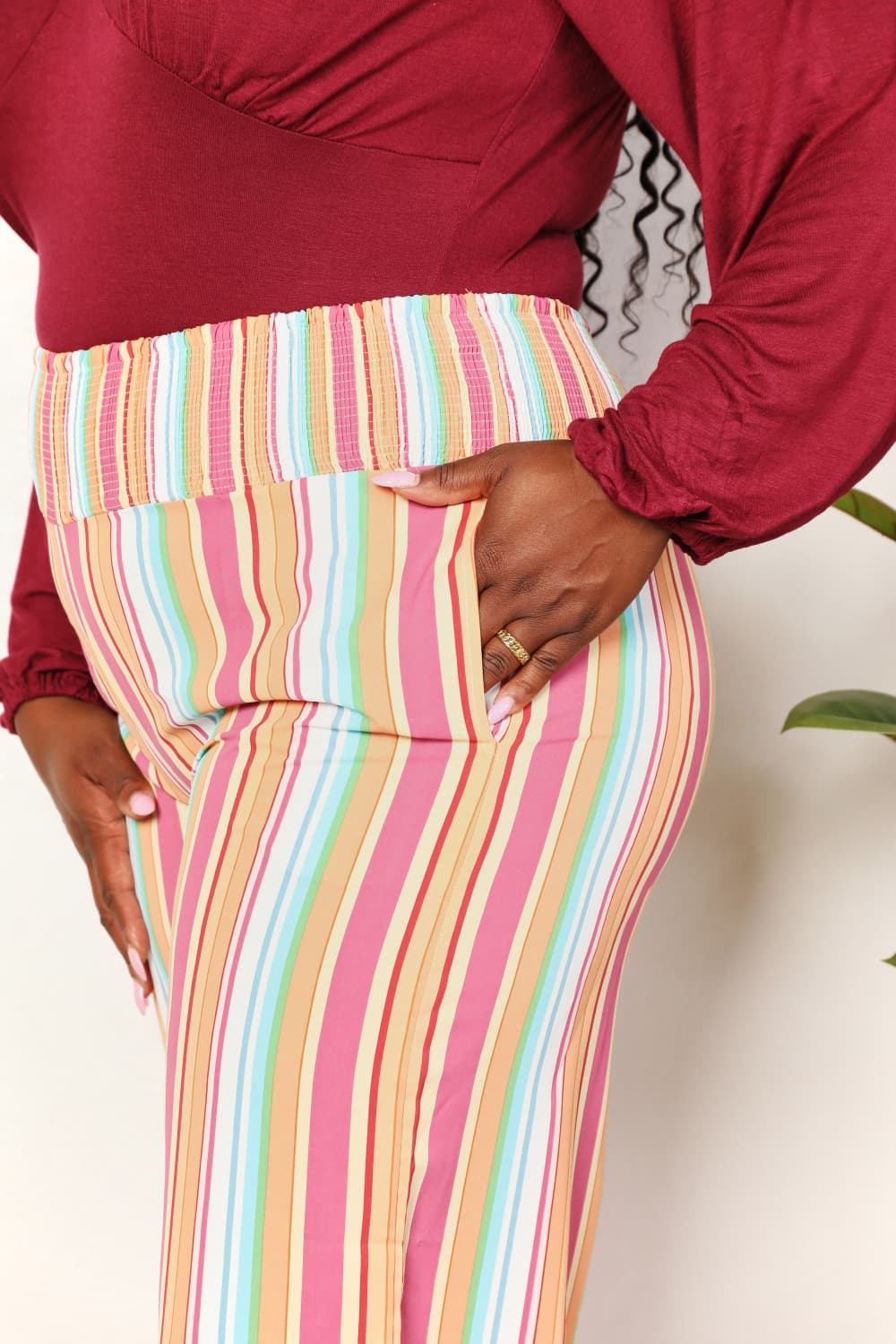 A person wearing Double Take Striped Smocked Waist Pants with Pockets and a burgundy long-sleeve shirt stands beside a wooden stool with a hat on it.