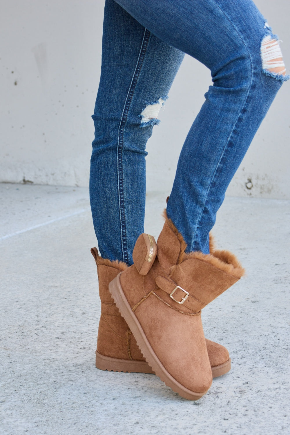 Person wearing ripped blue jeans and Forever Link Plush Thermal Flat Boots, tan in color, fur-lined with side buckles, standing on a concrete surface. These stylish boots promise cozy insulation for the feet.