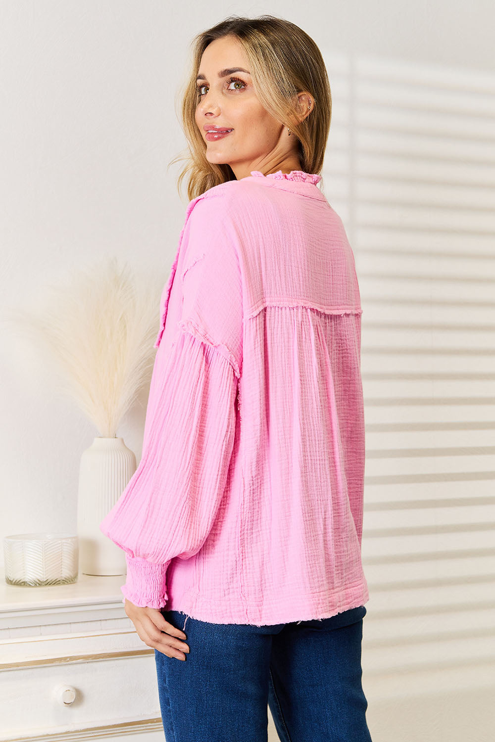 A person wearing a Double Take Exposed Seam Buttoned Notched Neck Blouse in pink and blue jeans touches their ear while standing indoors.