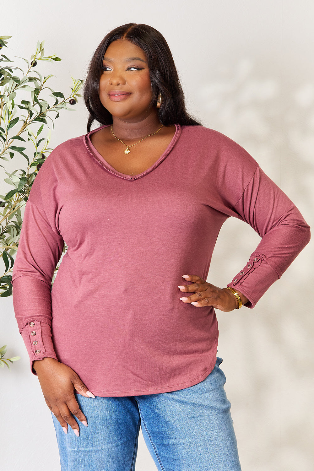 A person stands in front of a plain backdrop with a plant, wearing a maroon Culture Code Full Size V-Neck Exposed Seam Long Sleeve Blouse and blue jeans, smiling slightly with their hand on their hip. The relaxed fit of the polyester blend adds to the casual yet stylish look.