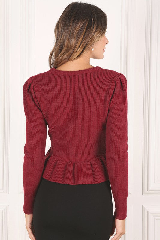 A woman stands elegantly in front of a white panel background, wearing the Peplum sweater top in red, featuring a round neck and puff sleeves, paired with a black skirt that has a side slit.