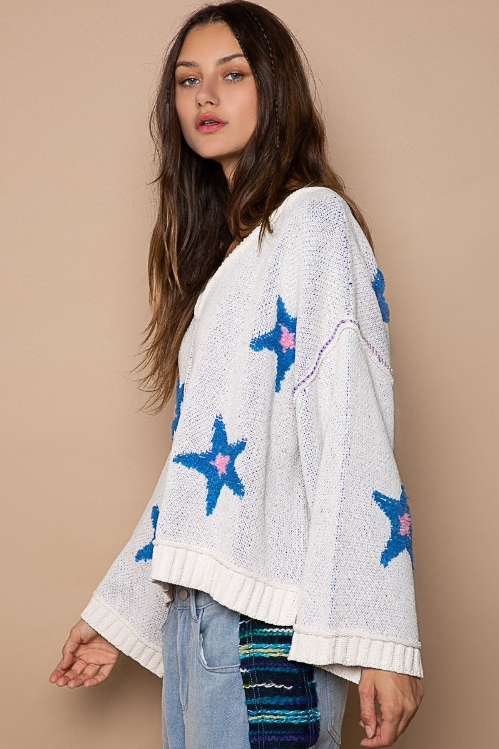 Woman in a POL Long Sleeve Star Patch Sweater with blue color-blocked star berber patches and light blue jeans standing against a beige background.