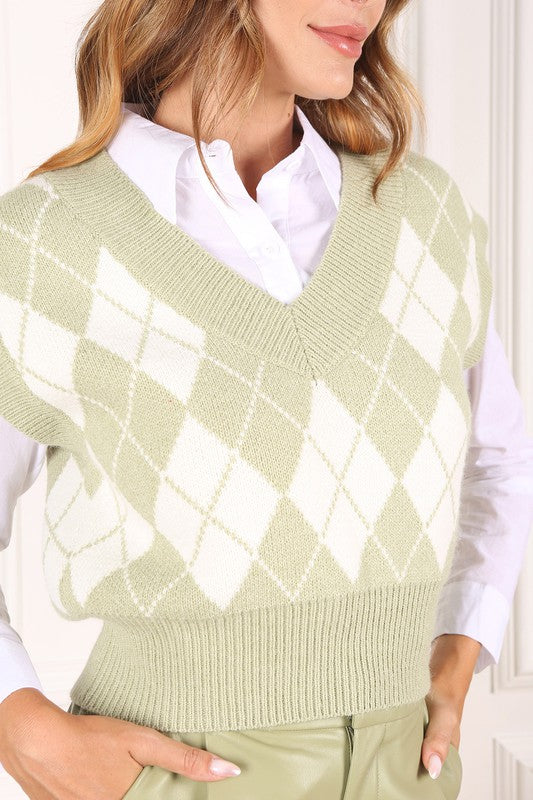 A person wearing a knitted argyle sweater vest in light green over a white shirt stands against a neutral background.