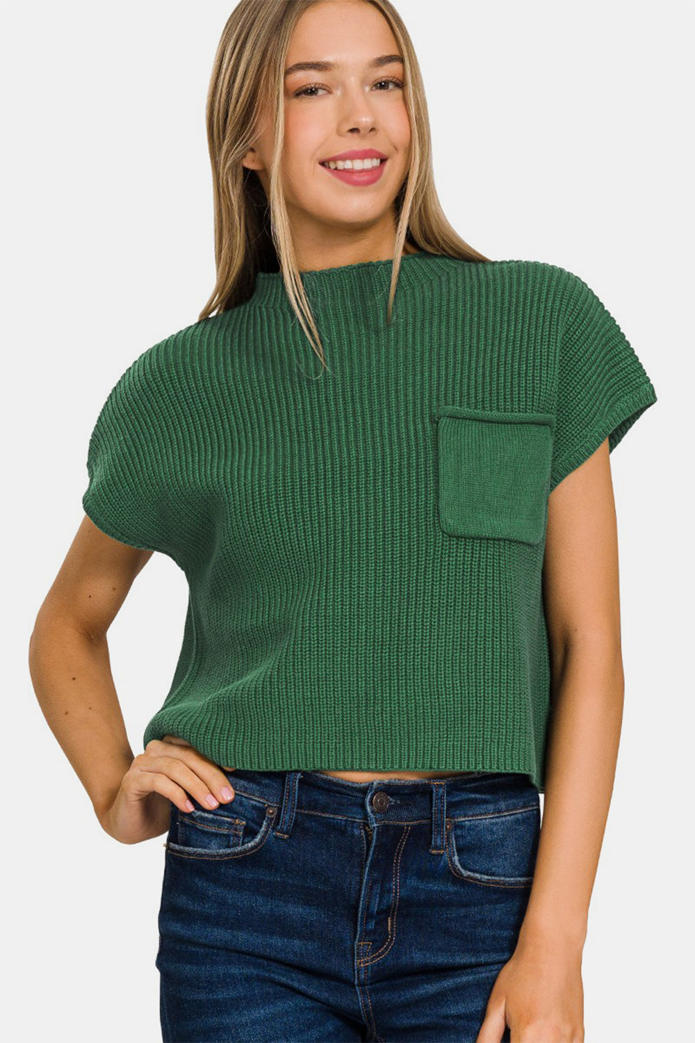 A woman wearing the Zenana Mock Neck Short Sleeve Cropped Sweater in green, featuring a small front pocket and crafted from knit fabric, is standing and smiling. She has long, straight hair and is paired with dark blue jeans.