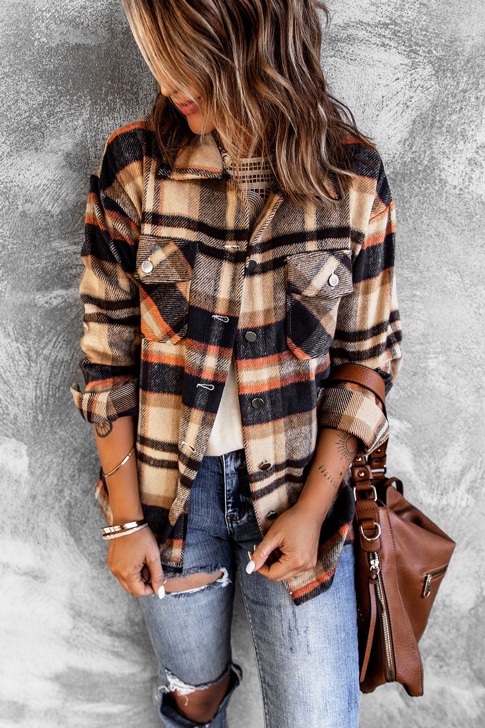 A person wearing a Brown Plaid Button Up Long Sleeve Flannel Shacket layered over a white top with ripped jeans and holding a brown handbag stands against a textured gray wall.