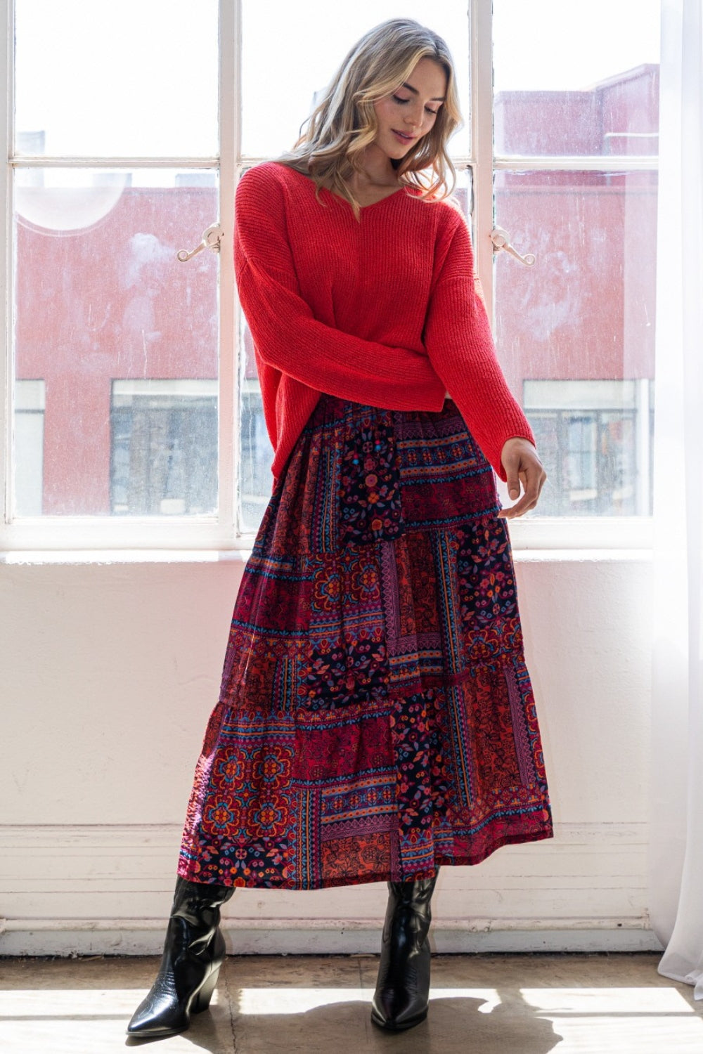 A person stands near a window exuding bohemian charm in a red sweater, an ODDI Full Size Printed Tiered Maxi Skirt, and black boots.