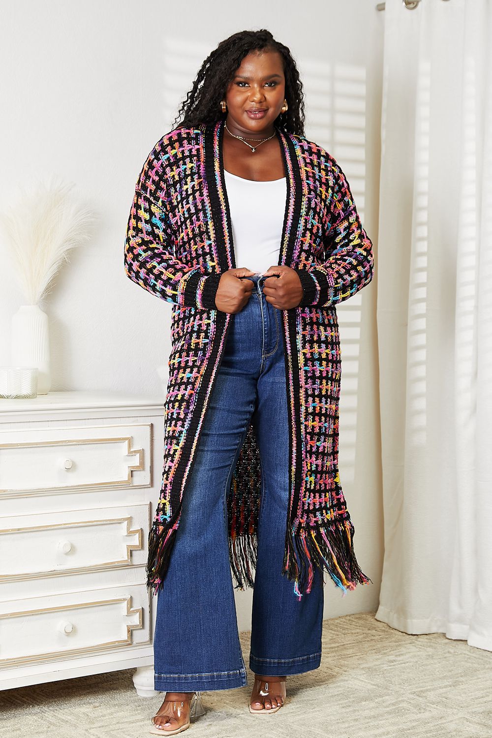 Person stands indoors wearing the Angel Wings Full Size Multicolored Open Front Fringe Hem Cardigan with a bohemian style over a white top and blue flared jeans.