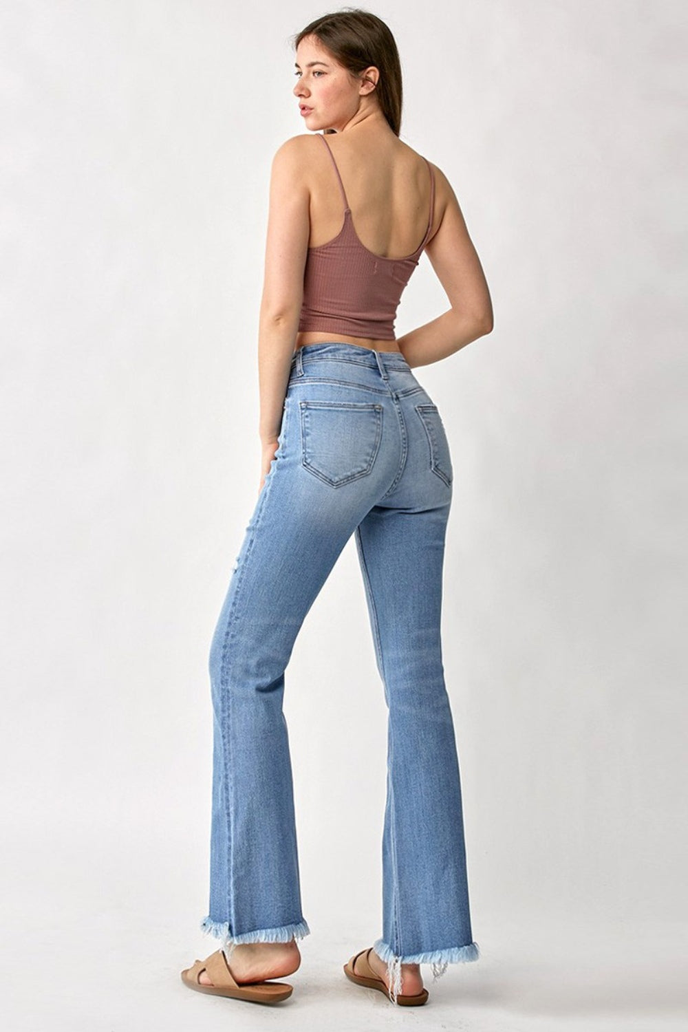 A person wearing RISEN High Rise Frayed Hem Bootcut Jeans in light-blue stretchy denim paired with tan sandals, standing against a plain background.