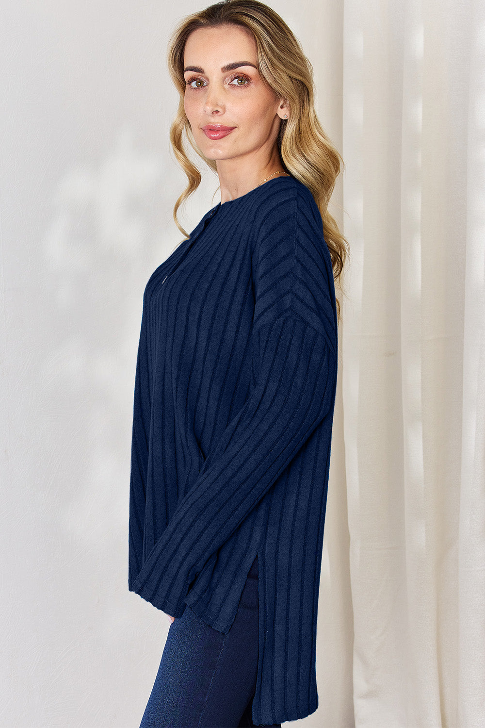 A person with long blonde hair is standing against a light background, wearing the Basic Bae Full Size Ribbed Half Button Long Sleeve High-Low T-Shirt in blue and dark pants. They are looking at the camera and smiling slightly, showcasing the chic high-low hem of their top.