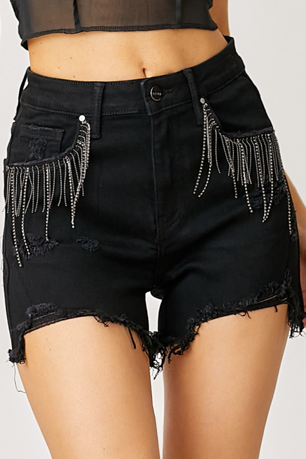 A person wearing RISEN Frayed Hem Denim Shorts with Fringe Detail Pockets, featuring decorative chains hanging from the front pockets, perfect for summer fashion. The high-waisted shorts have a distressed look with a frayed hem and fringe detail pockets for an edgy style.