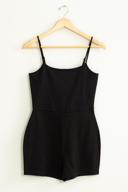 The Loving Me Fitted Sleeveless Romper, featuring a fitted bodice and spaghetti straps in black, hangs gracefully on a wooden hanger against a white wall.