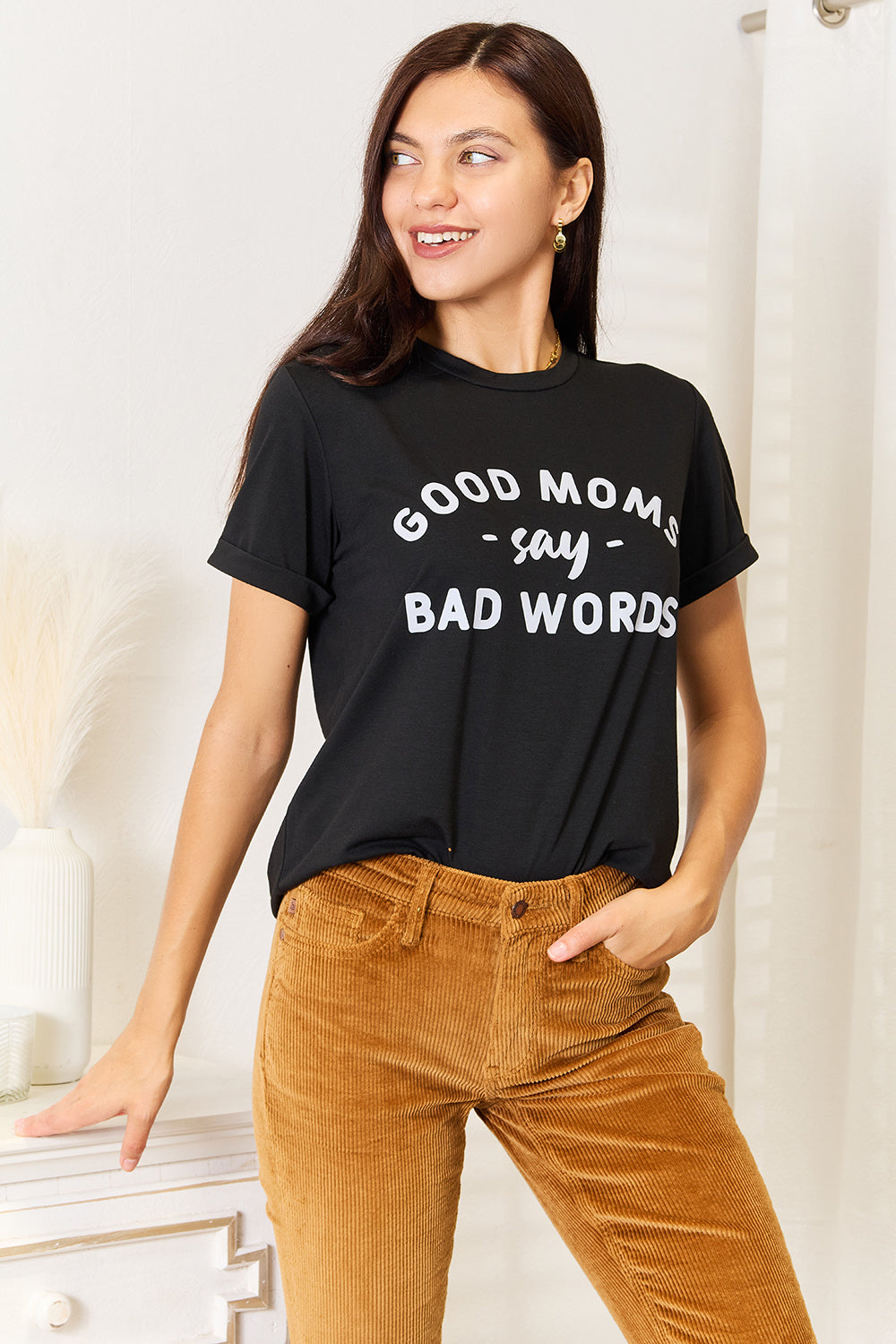 A woman stands in a room with a white wall, where light filters through the blinds, subtly showcasing her rebellious side while wearing the Simply Love GOOD MOMS SAY BAD WORDS Graphic Tee.