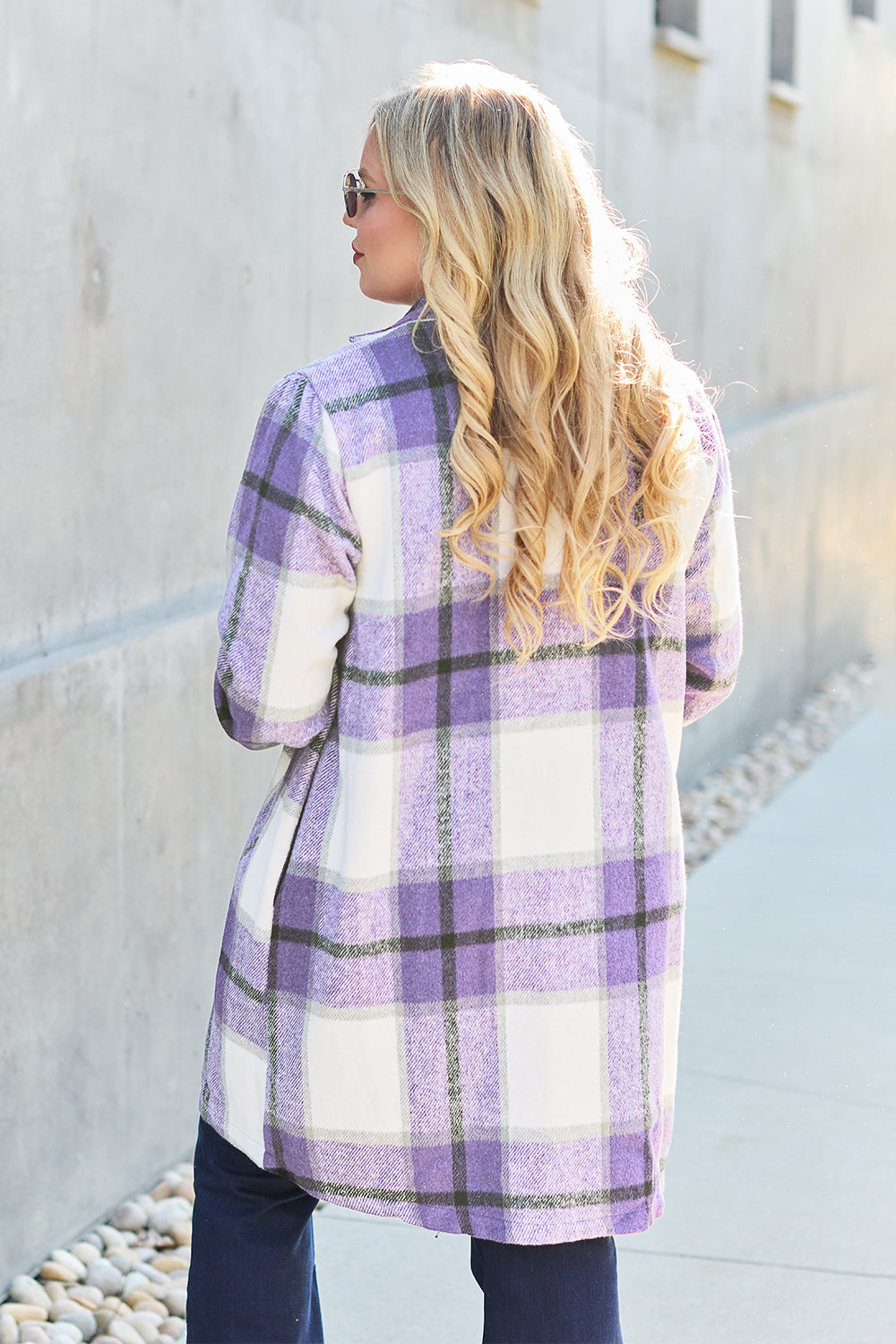 A woman stands on a sidewalk wearing the Double Take Full Size Plaid Button Up Lapel Collar Coat in pink and white over a white top and blue jeans, with her right hand lightly touching her hair. The polyester fabric of the coat makes it durable and perfect for an easy machine wash cold.