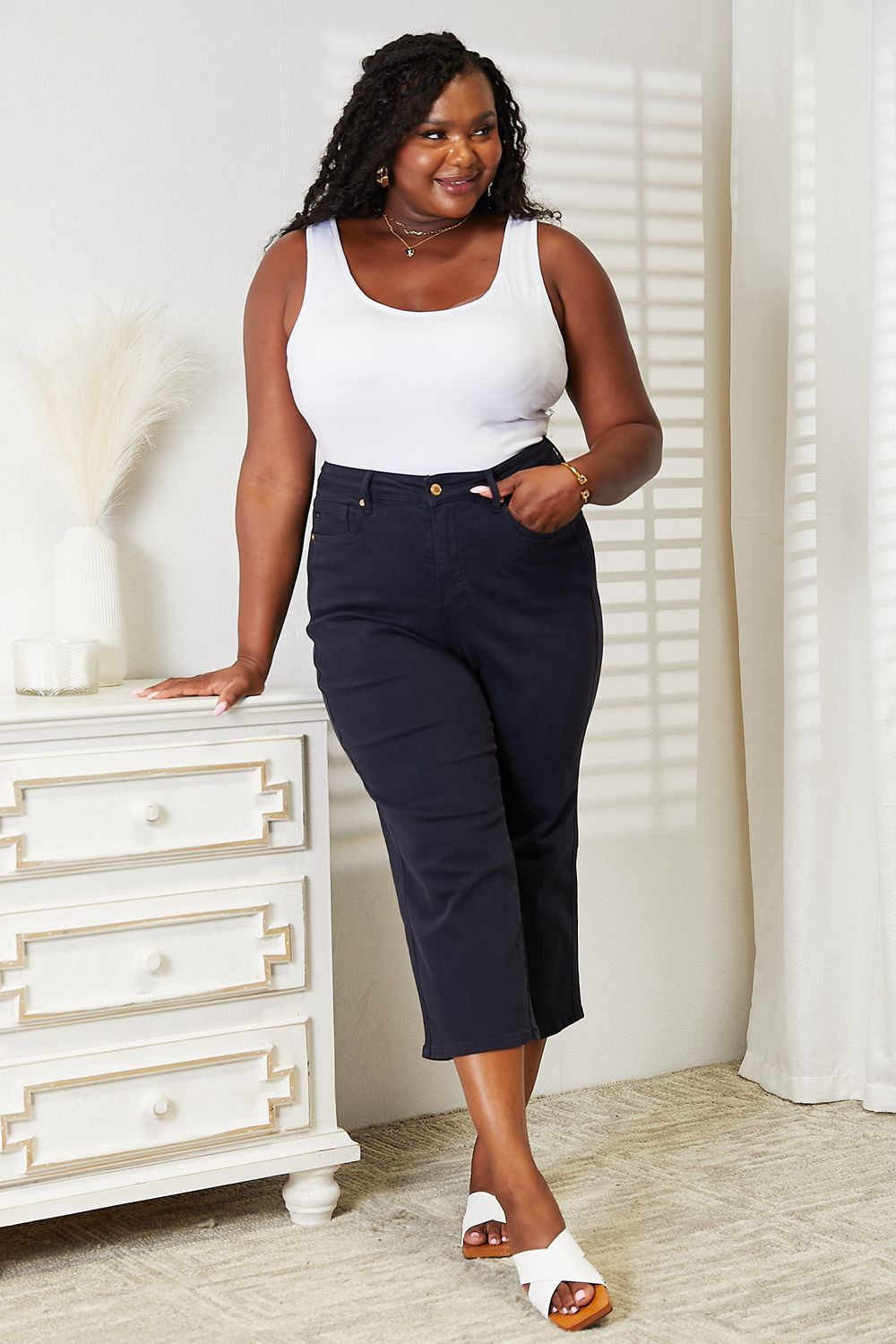 A person wearing a white sleeveless top and Judy Blue Full Size High Waist Tummy Control garment-dyed wide cropped jeans in black stands indoors next to a white dresser.