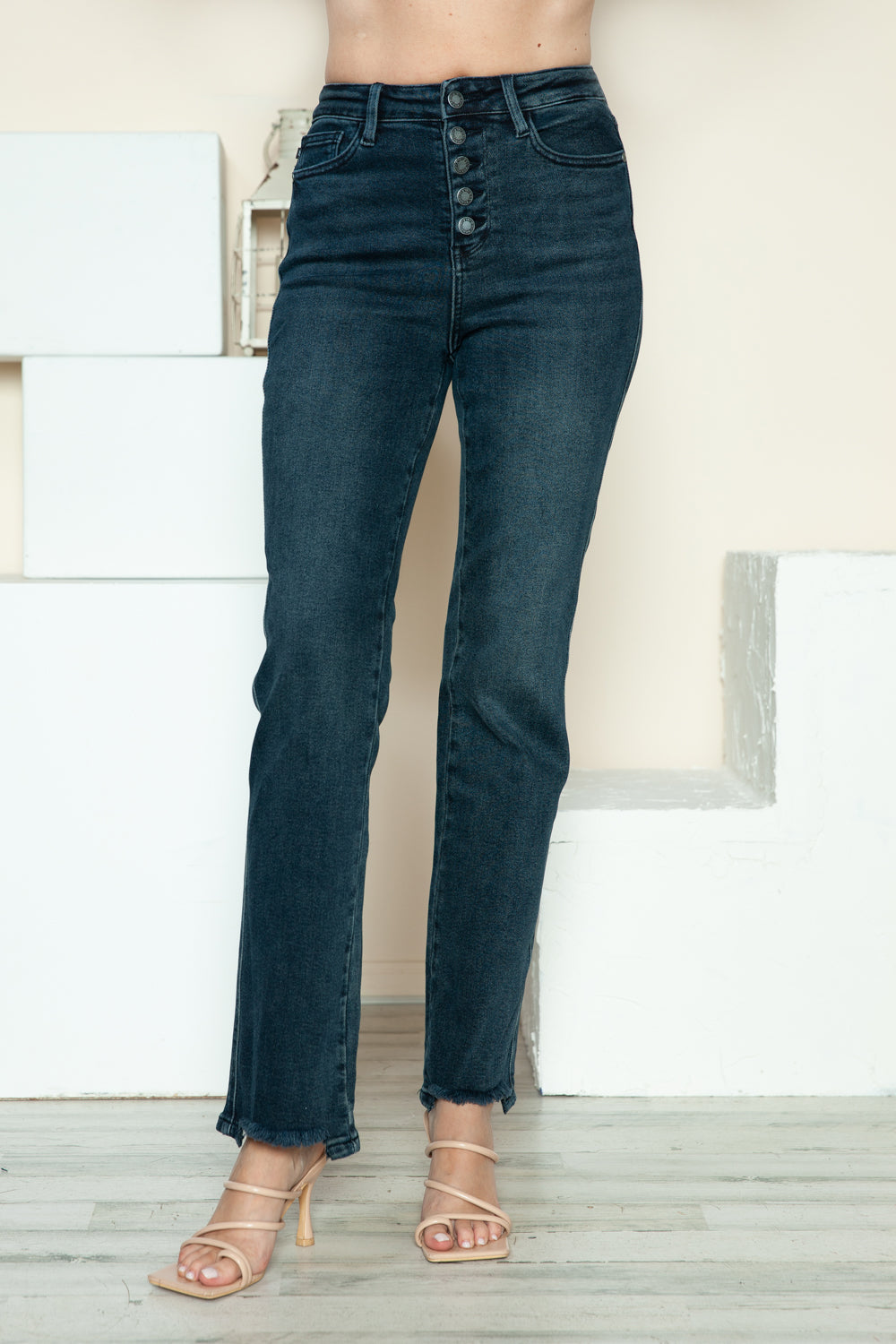 A person is modeling the Judy Blue Full Size Button Fly Hem Destroy Straight Jeans, high-waisted with distressed detailing and frayed hems, paired with light-colored open-toe heels. The background includes white blocks and a light-colored wall.