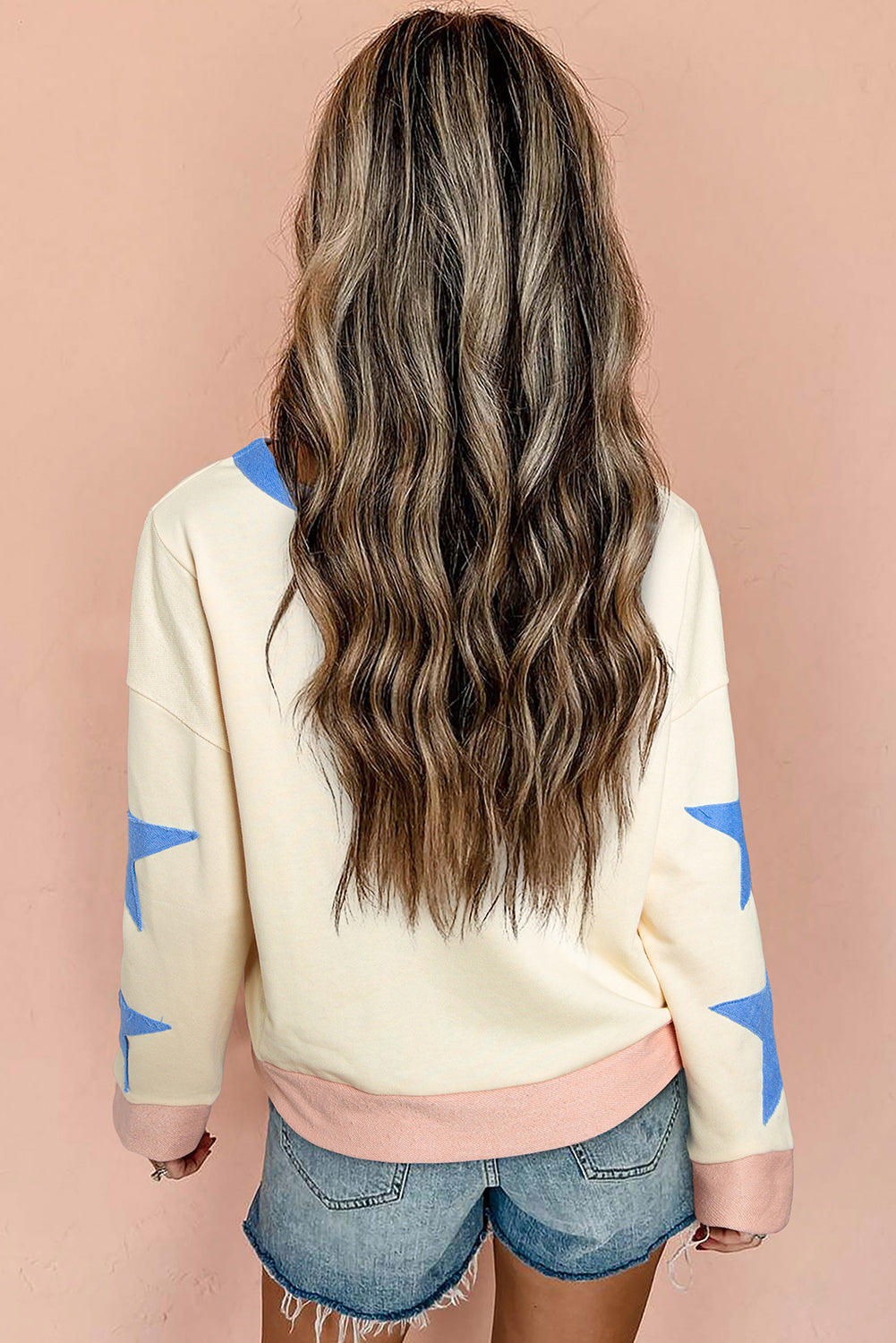 A person with long, wavy, light brown hair stands facing away, wearing the Beige Star Patchwork Exposed Seam Oversized Sweatshirt paired with denim shorts. The background is a plain, light peach wall, adding an edgy vibe to the look.