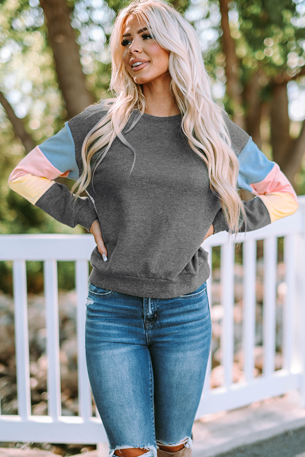 A person with long blonde hair wearing a stylish Gray Colorblock Long Sleeve Pullover Sweatshirt and blue jeans stands outdoors near a white fence.