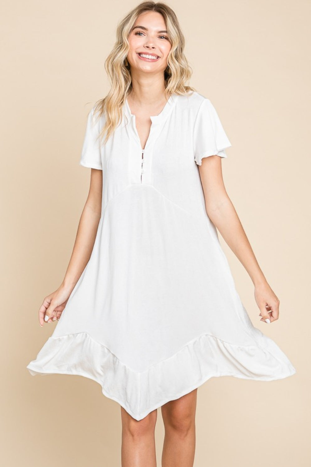 A person wearing the versatile white Culture Code Full Size Short Sleeve Ruffled Asymmetric Hem Dress stands in front of a plain beige background, showcasing its flattering fit.