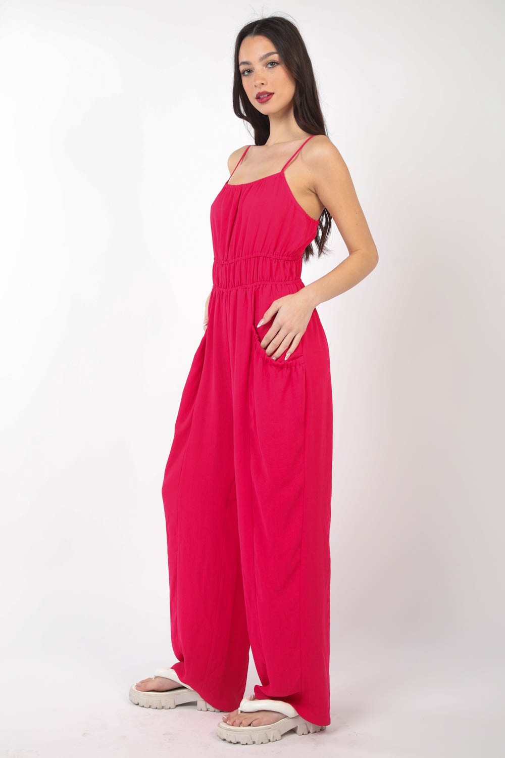 A person wearing the VERY J Pintuck Detail Woven Sleeveless Jumpsuit in bright pink paired with white sandals stands against a white background, with one hand placed in a pocket.