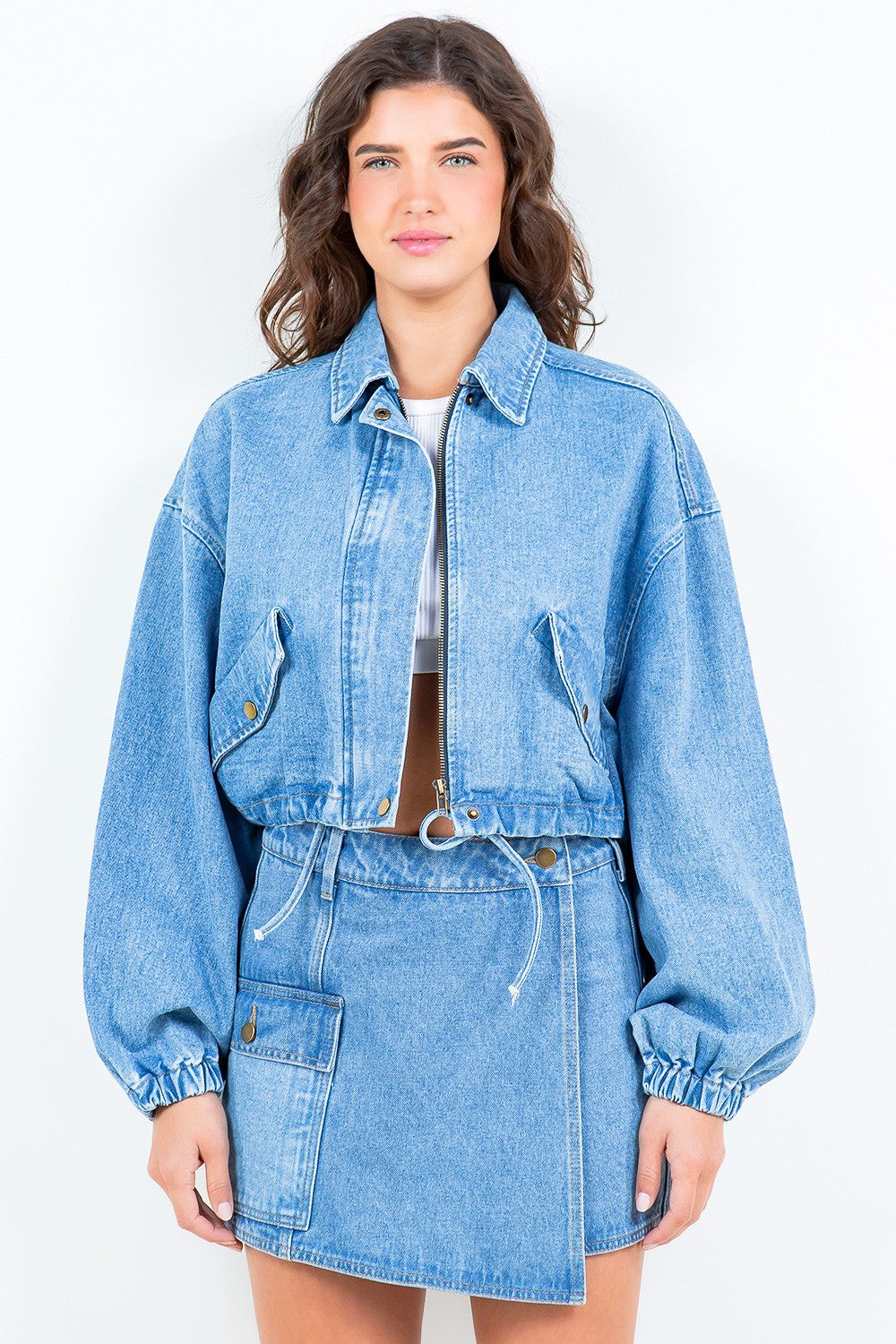 A woman wearing the American Bazi Drawstring Waist Crop Denim Jacket in light blue denim and a matching skirt, standing against a plain light background.