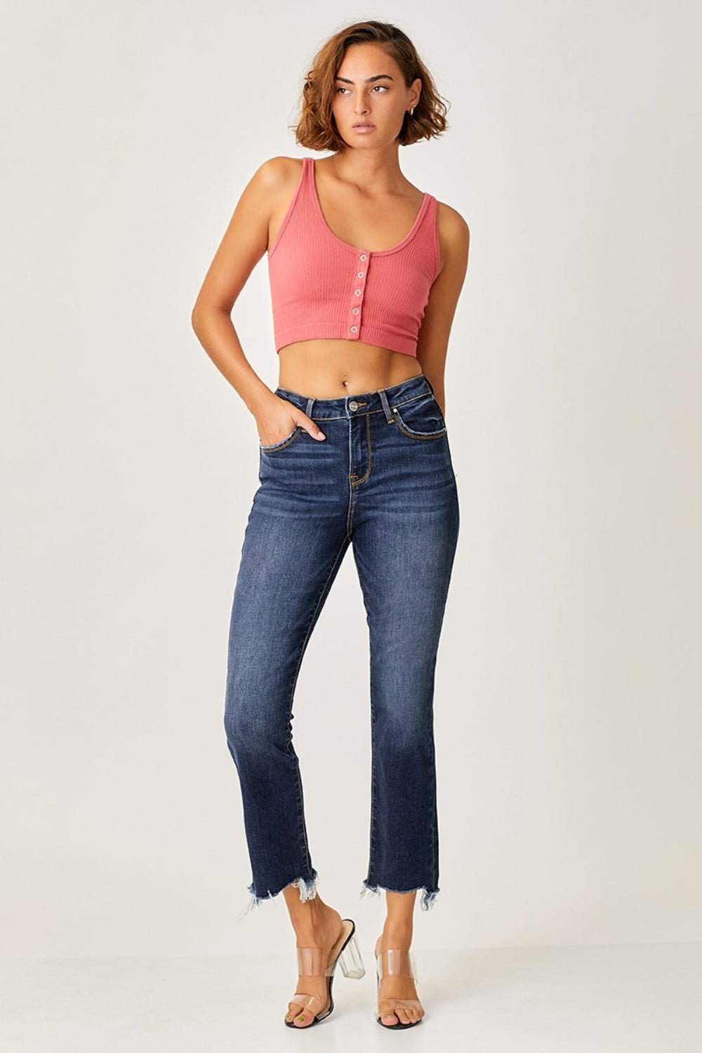 A person striking a pose in Risen Full Size Frayed Hem Cropped Straight Jeans with clear high-heeled sandals, one hand casually tucked in a pocket against a plain background.