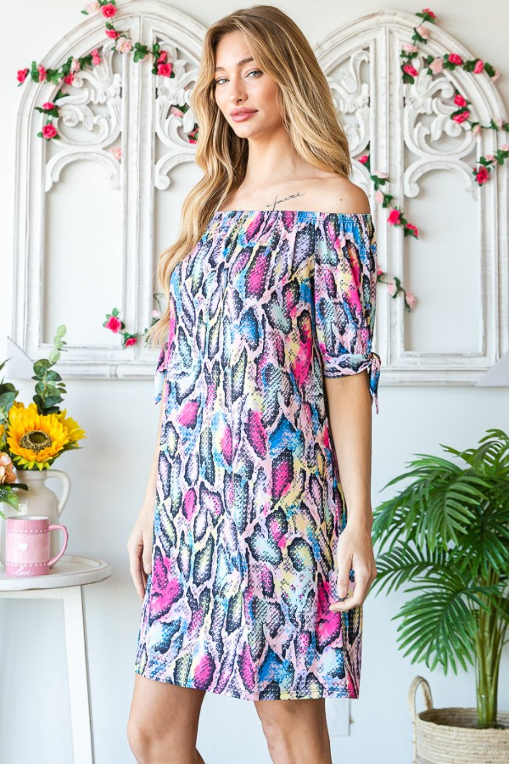 A woman in a stylish Heimish Full Size Off Shoulder Snake Mini Dress and a large straw hat stands next to indoor plants.