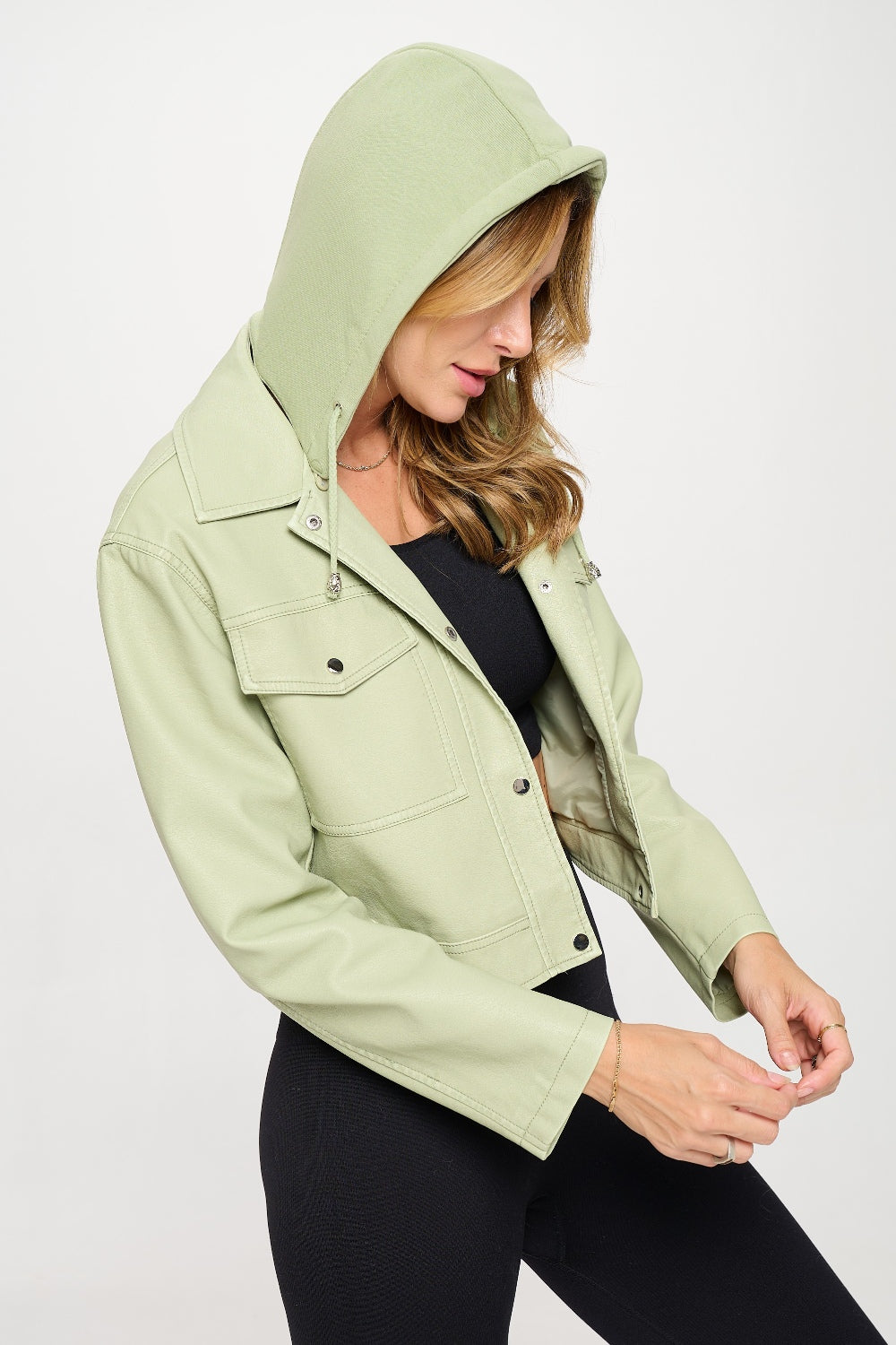 A person showcases a trendy Coalition LA Snap Down Cropped Hooded Jacket in light green over a sleek black outfit against a plain background.